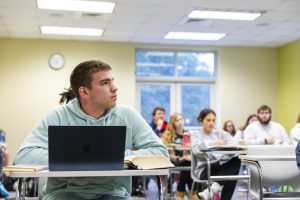 Male student in light blue jacket sitting in a desk in the forefront with a computer. Other students are behind him in their desks.